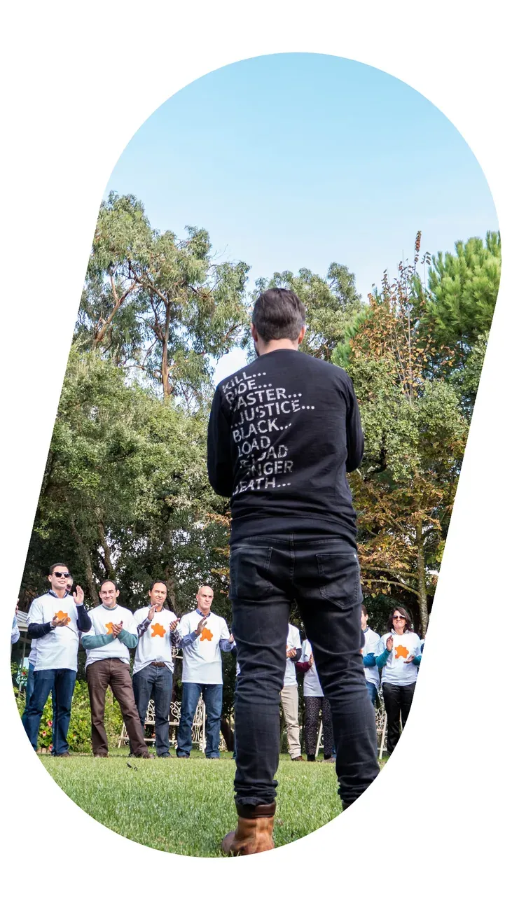 A team in white shirts in a semicircle on the grass, listening to a facilitator.