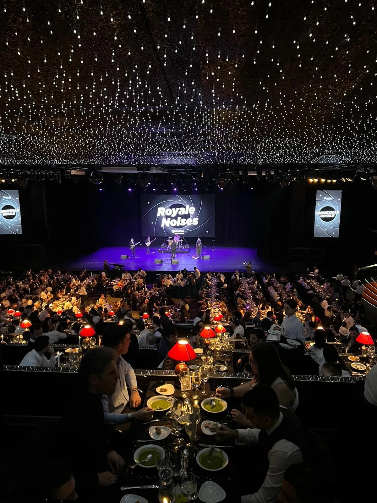 A gala dinner in a dark room, ceiling strung with lights, round tables full and a stage with two screens.