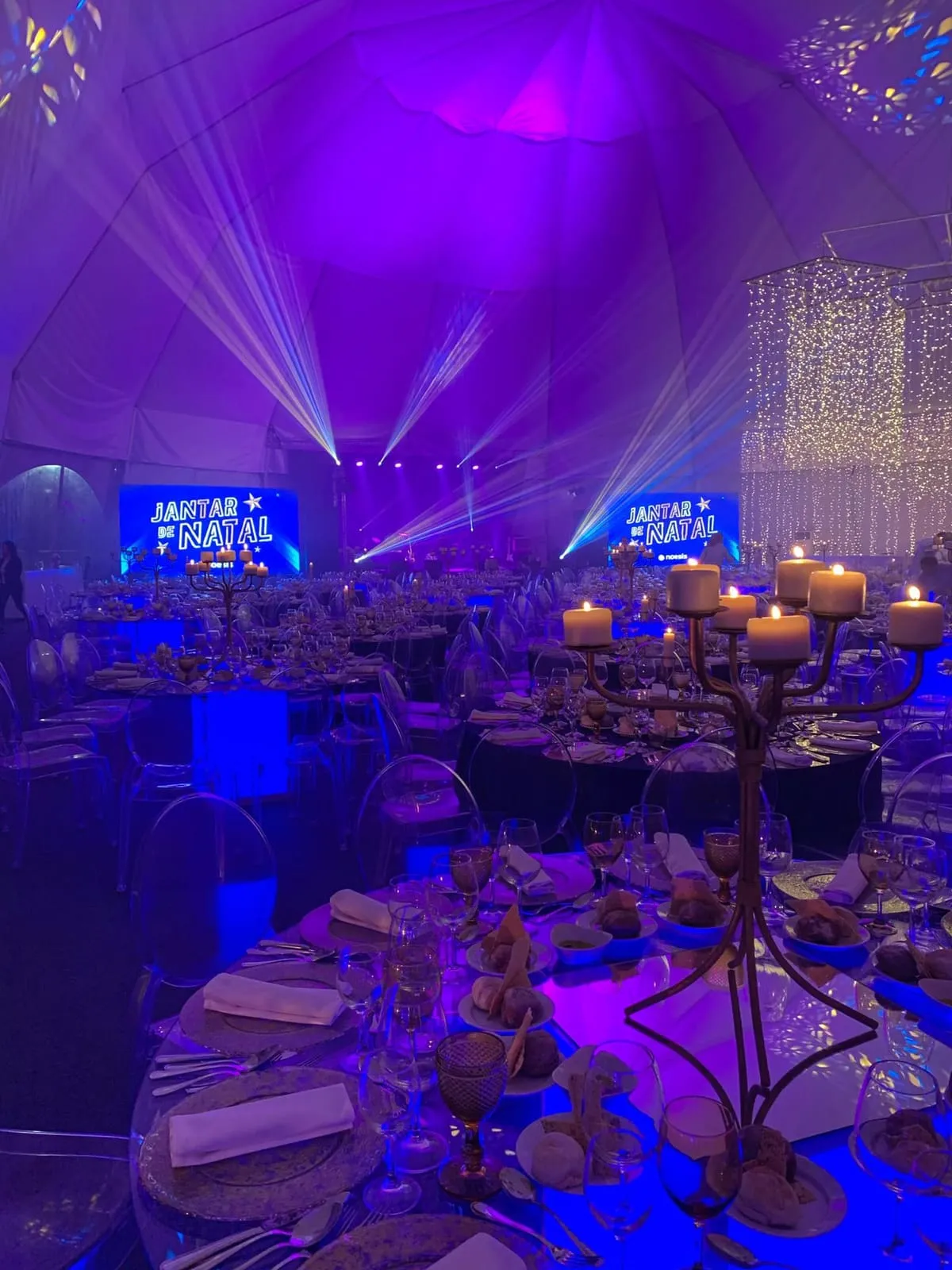 A hall lit in purple and blue, tall candelabra on the tables and beams of light over the floor.