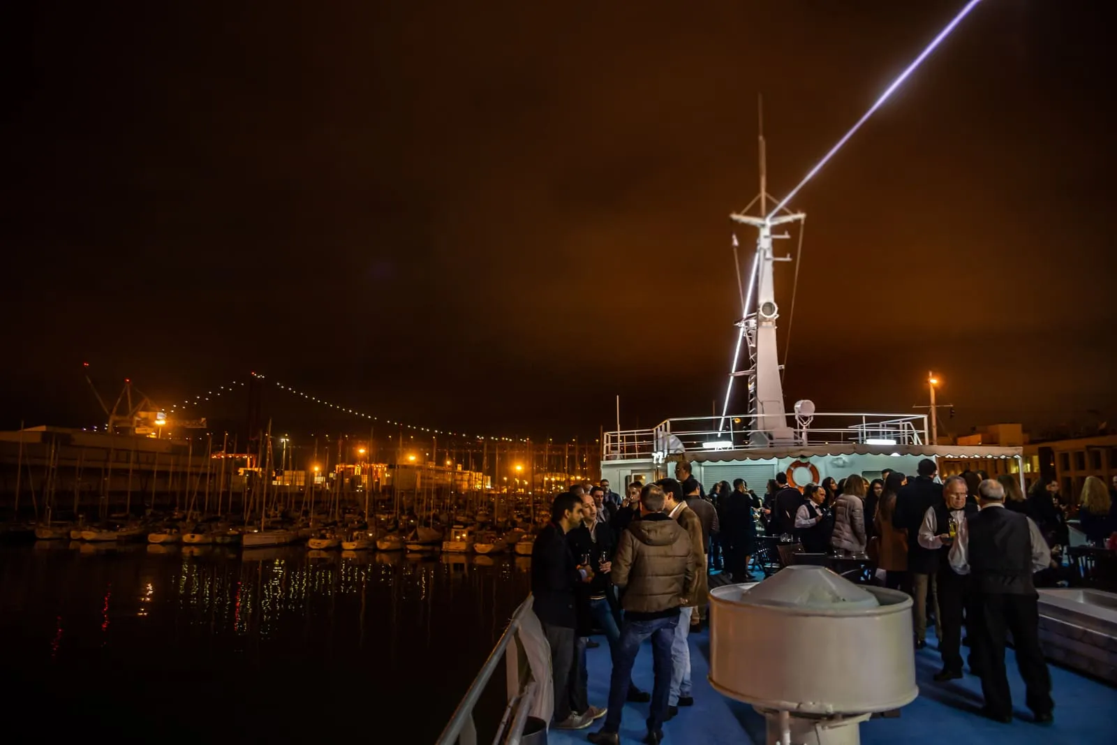 The deck of a boat at night, guests standing, city and bridge lights behind.
