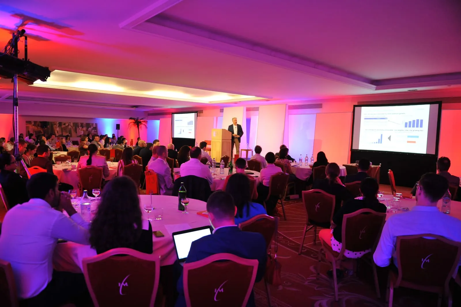 A convention dinner lit in pink, long tables and a presentation running on screen.