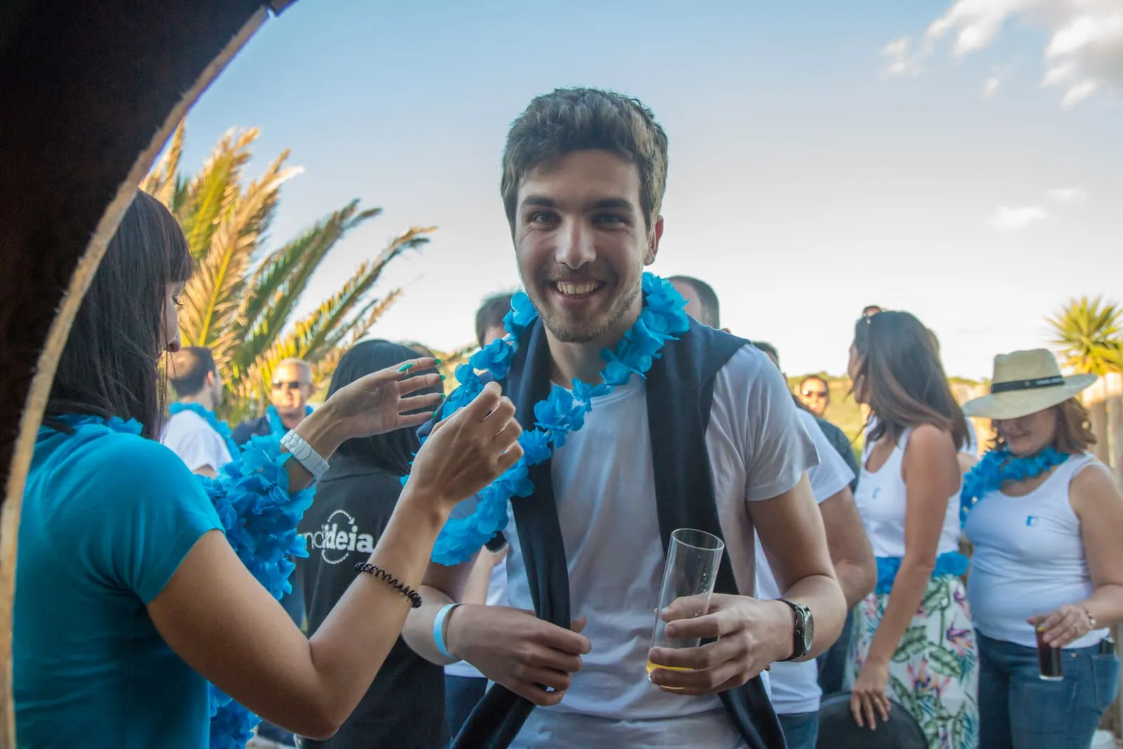 Participantes com lenços azuis ao pescoço, de copo na mão, num evento ao ar livre.