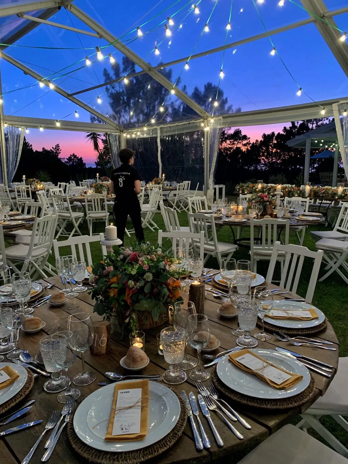 A clear marquee at dusk, fairy lights overhead and tables set waiting for guests.