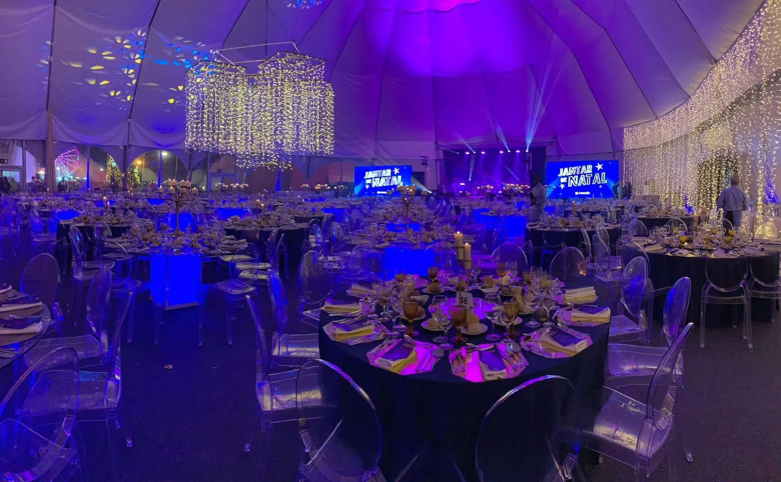 Jantar de gala numa tenda iluminada de roxo e azul, com mesas redondas postas a ouro, cadeiras transparentes e dois ecrãs no palco.