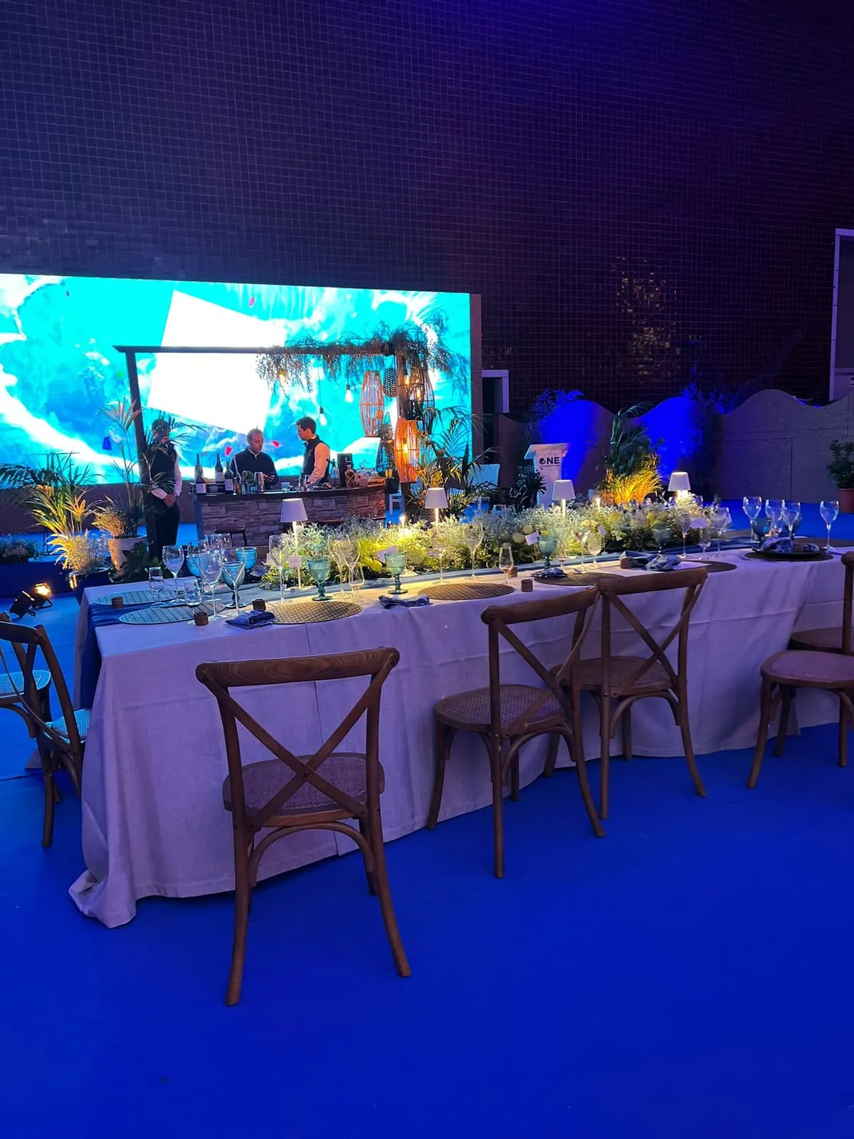 Mesa comprida posta a branco, com cadeiras de madeira e arranjos baixos de flores, diante de um ecrã gigante com uma paisagem tropical.