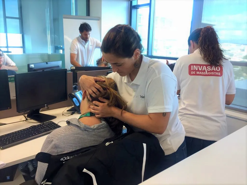 Massagistas de camisola branca com o dizer "Invasão de Massagistas" a massajar colegas sentados nas suas secretárias, junto às janelas do escritório.