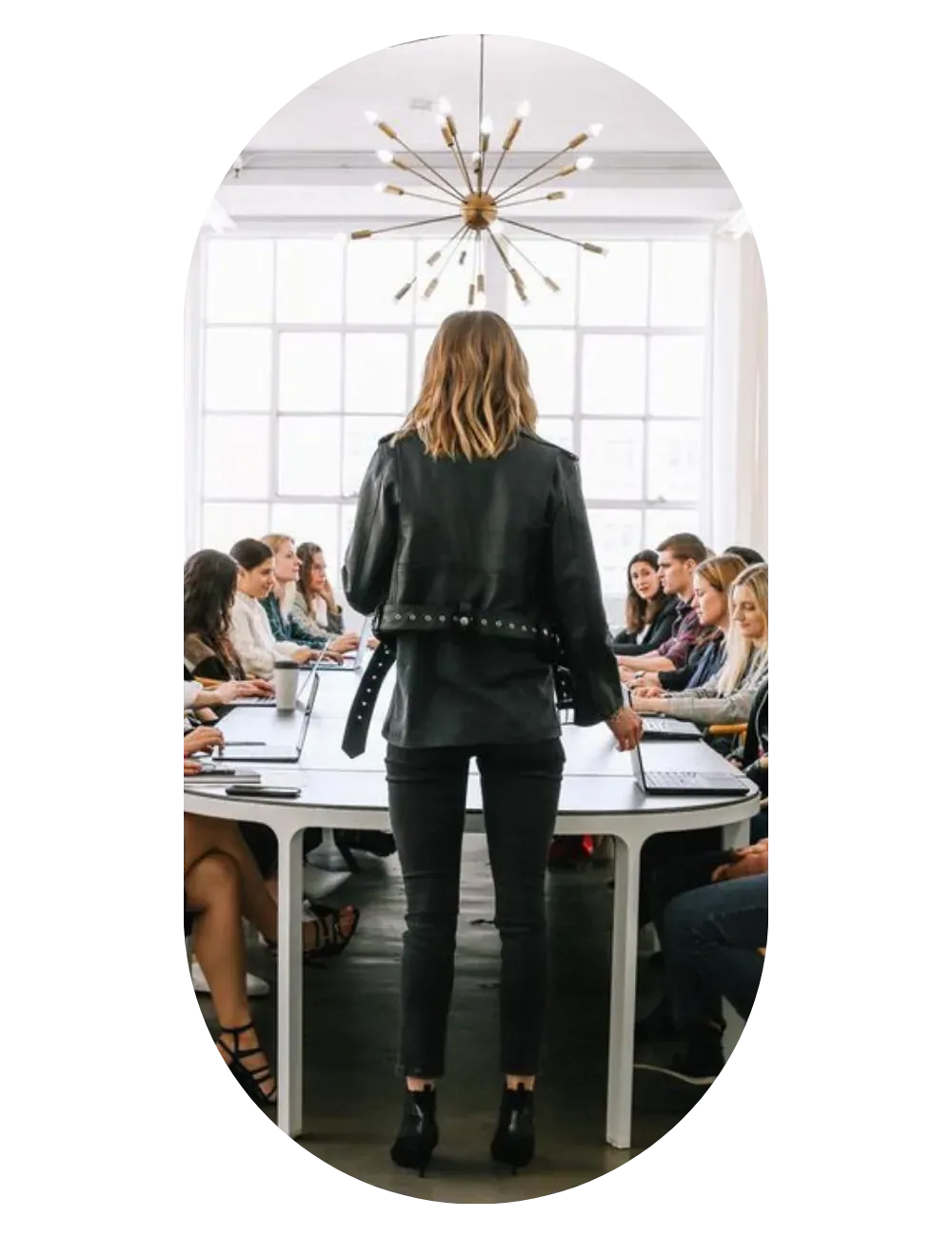 A person standing on a long table addressing the team seated around it, in a bright room.