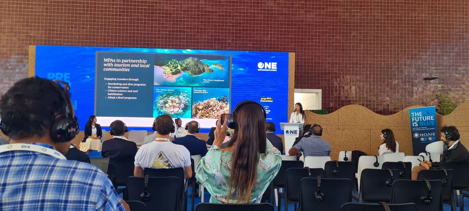 Audience watching a talk about the ocean, with the ONE logo on screen.