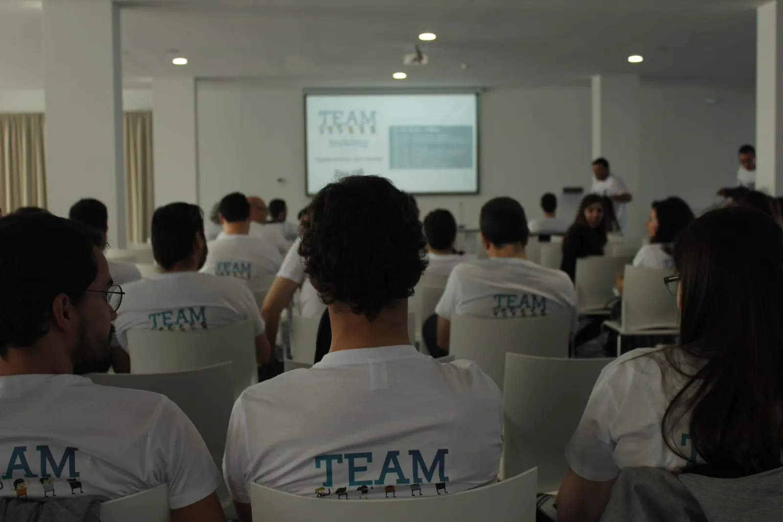 Participants in team t-shirts, seated and listening to a presentation.
